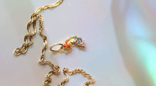 A multi-coloured rainbow of sapphire gemstones - red, orange, yellow, green, blue and pink - with a gold necklace chain and rainbow-lit background, celebrating pride